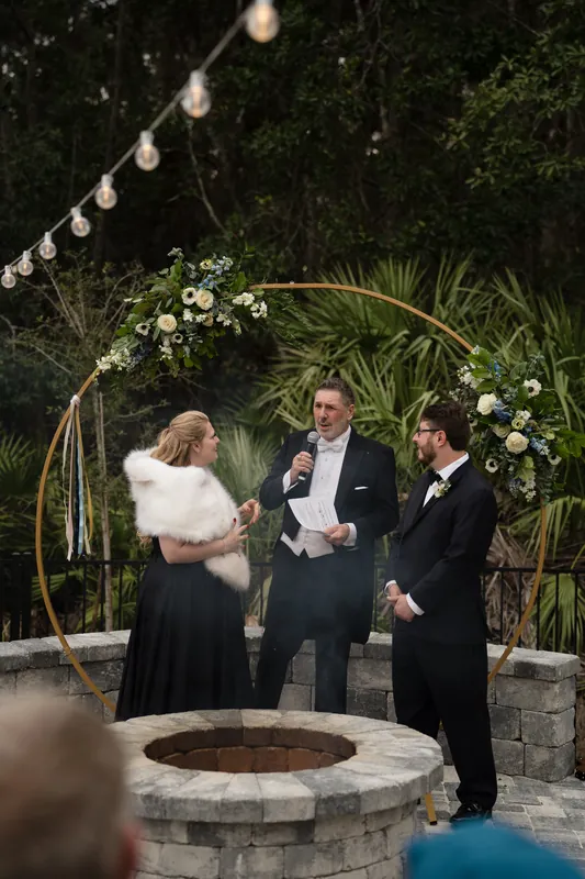 Three guests gather around a stone fire pit beneath hanging lights, with tropical greenery and a floral arch framing the evening scene.