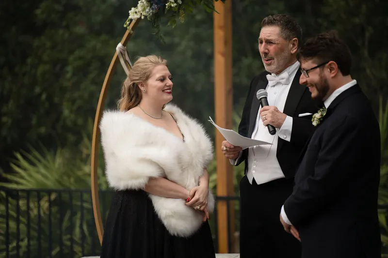 Someone in a cream gown and white wrap holds a bouquet while standing beside two men in dark suits near a wooden arch.