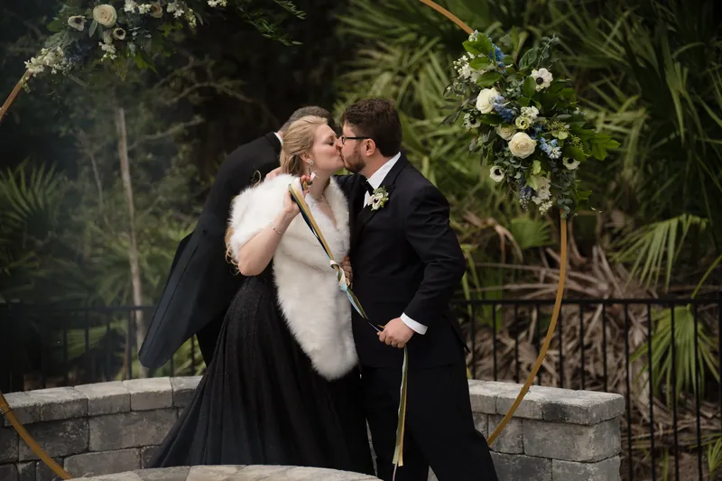 Two people in formal wedding attire kiss beside a floral arrangement, tropical plants and a decorative railing softly visible behind them.
