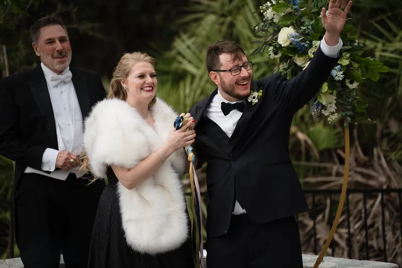 Two people in formal black attire celebrate with raised arms amid tropical greenery and floral décor.