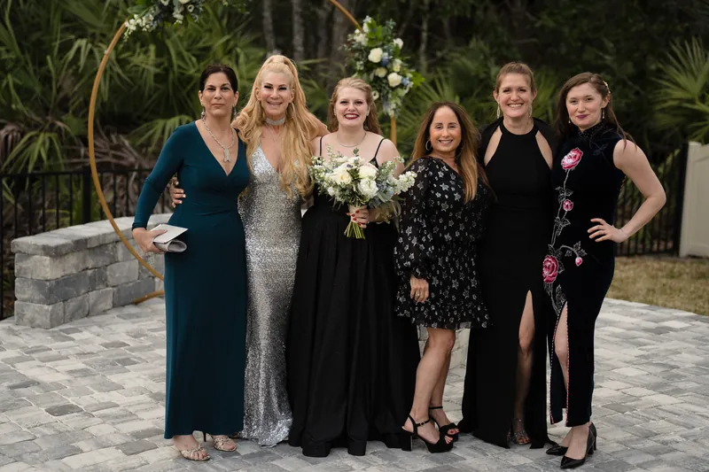 Six women in varied formal wear pose together on a paved terrace, framed by lush palms and a white flowering arch.
