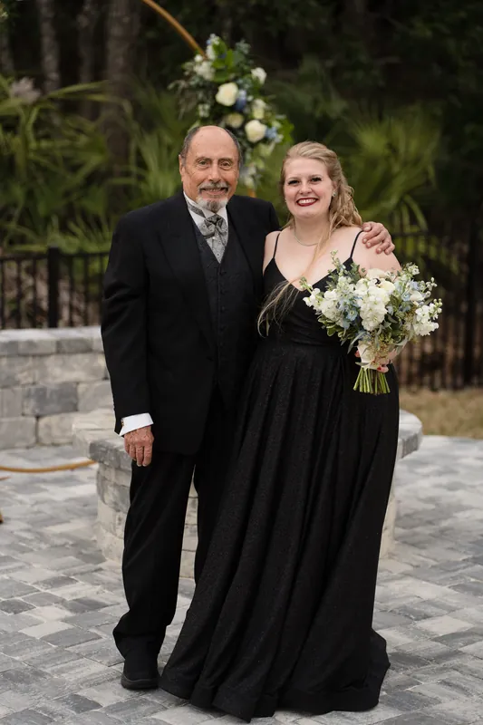 Two guests in black formal wear stand arm-in-arm holding a white bouquet, smiling at the camera with tropical foliage behind them.