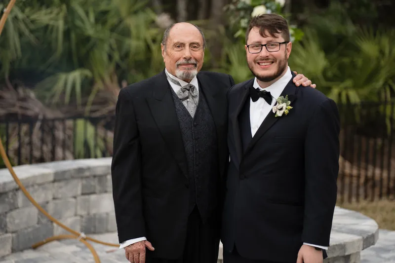 Two men in dark suits with boutonnières stand together with their arms linked, posing against a backdrop of greenery and stone steps.