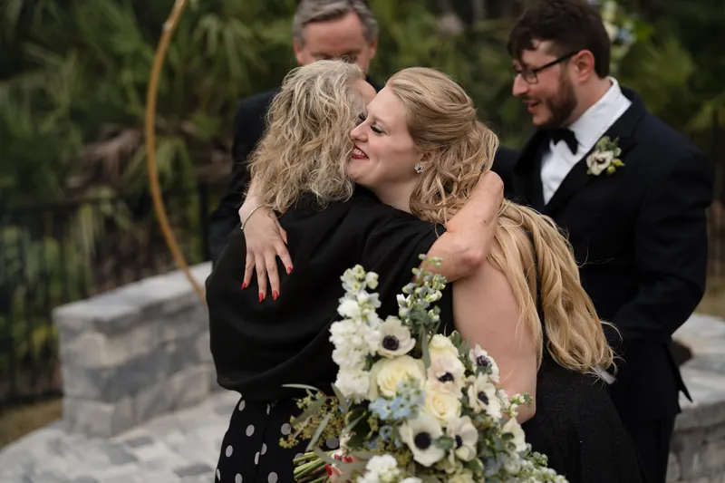 Four people embrace together outdoors, with someone in the center holding a white floral bouquet, surrounded by tropical vegetation.