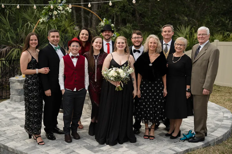 A group of eleven family members and guests gather on a stone patio, spanning three generations in formal attire and varying outfits.