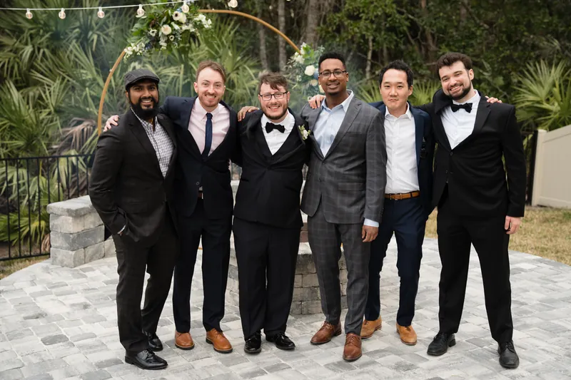 Six guests in formal black suits and ties stand shoulder-to-shoulder on a paved outdoor terrace lined with tropical plants.