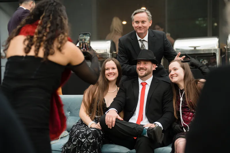 A small group socializes indoors, with someone in a red dress standing and others seated on a teal sofa in business attire.