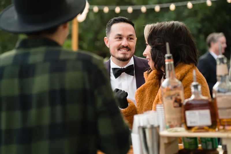 Someone in a tuxedo and bow tie converses with a guest in a gold cardigan at an outdoor reception with string lights overhead.