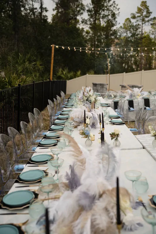 A long table set for dining stretches across a patio, with teal napkins, place settings, and pampas grass arrangements under evening lights.