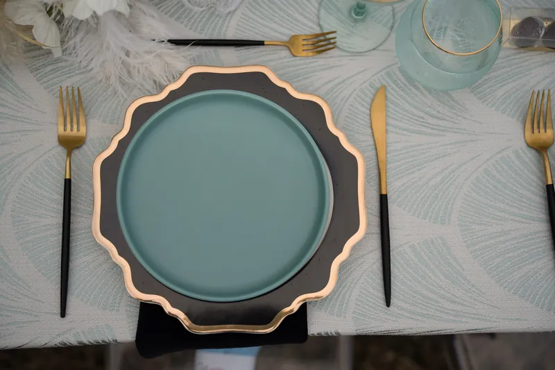 Place setting detail: teal scalloped plate, gold flatware, black napkin, and glassware arranged on a patterned gray tablecloth.