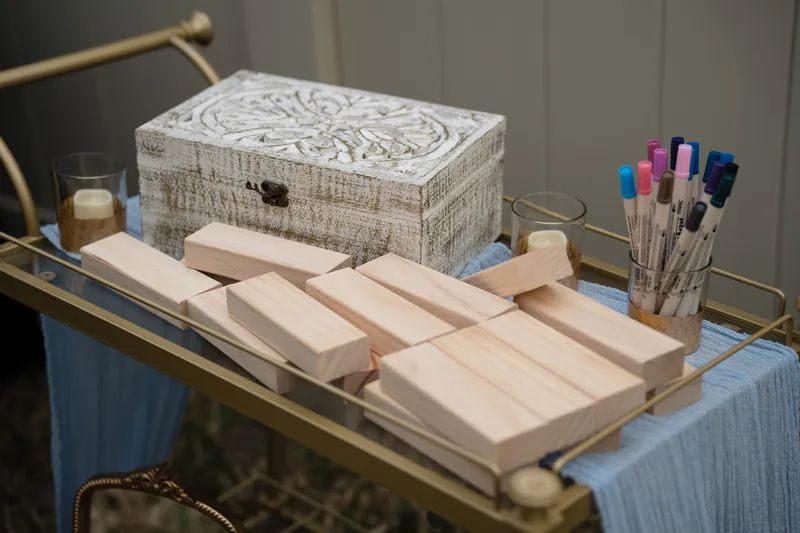 Blank wooden place cards and markers arranged on a gold table with a decorative white box and colored pens nearby.