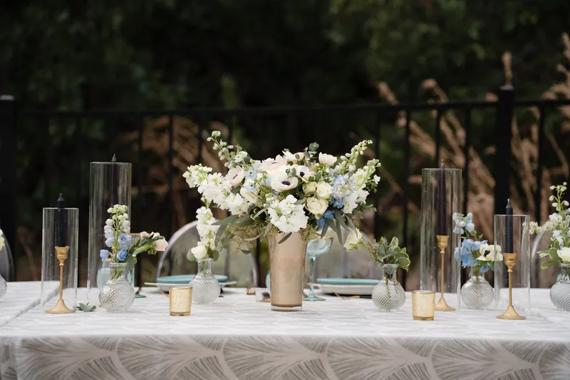 White anemone and greenery arrangements in tan vases line a long table set for an outdoor reception.