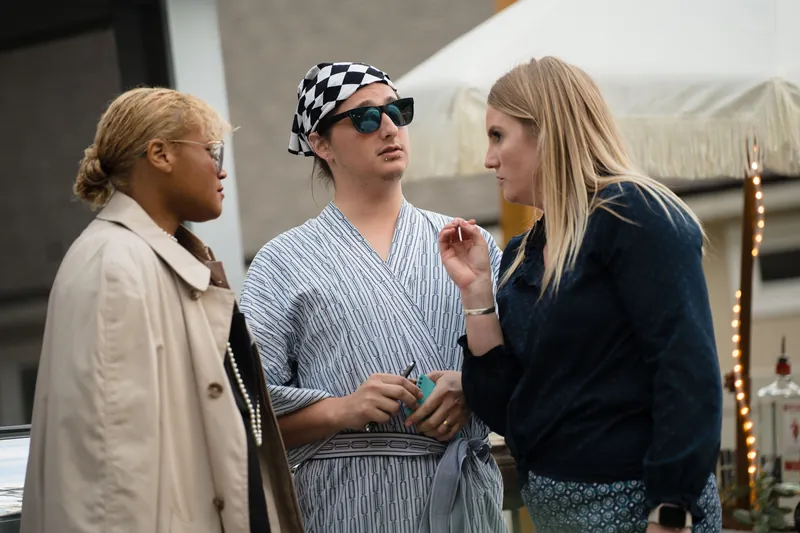 Three people stand together indoors, one wearing sunglasses and a patterned headwrap, chatting near warm lighting.