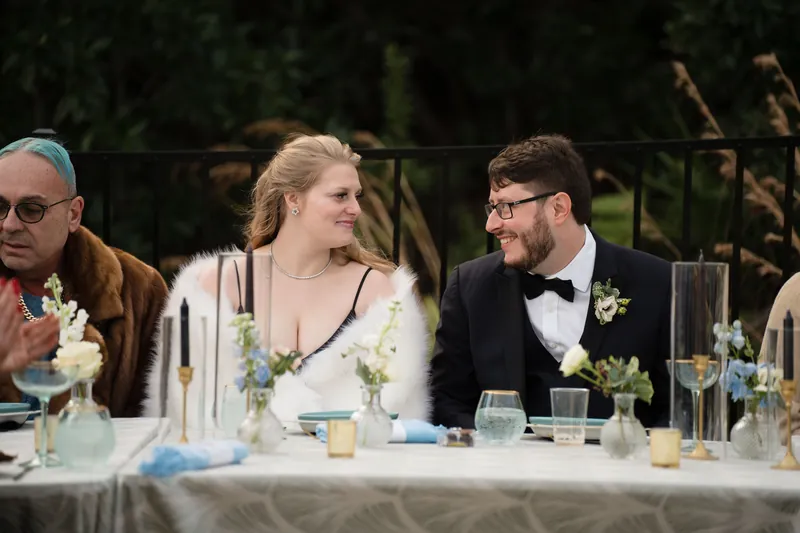 Four guests seated at an outdoor dining table smile and converse, with white flowers and glassware between them.
