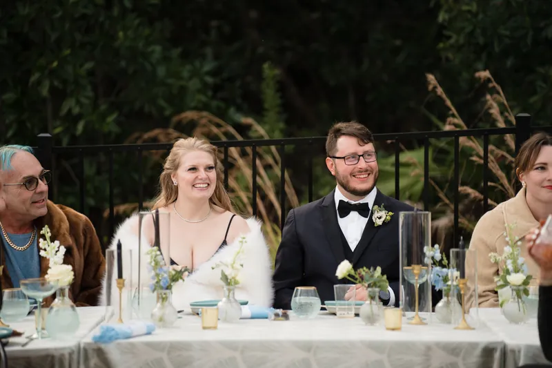 Five guests at an outdoor reception table engage in conversation, dressed formally with white floral arrangements and place settings visible.