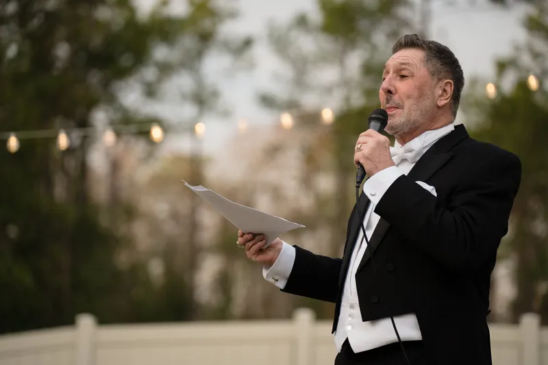 Person in formal attire holding a microphone and papers, speaking outdoors with blurred greenery behind.