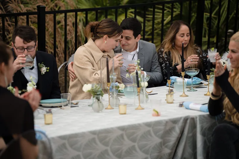 Six guests seated at an outdoor dining table with white linens, some holding microphones during speeches.