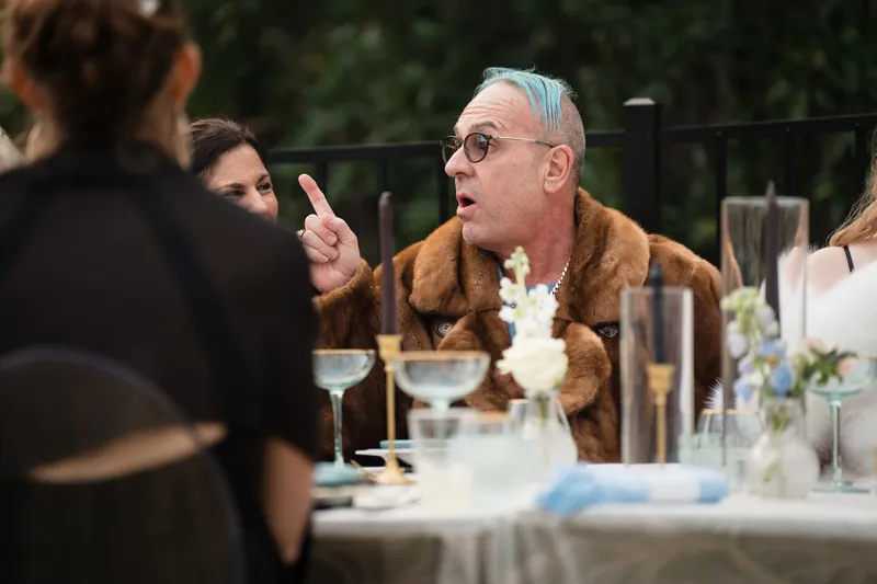 Older guest in tan cardigan gesturing animatedly while speaking at a dinner table lined with glasses and place settings.