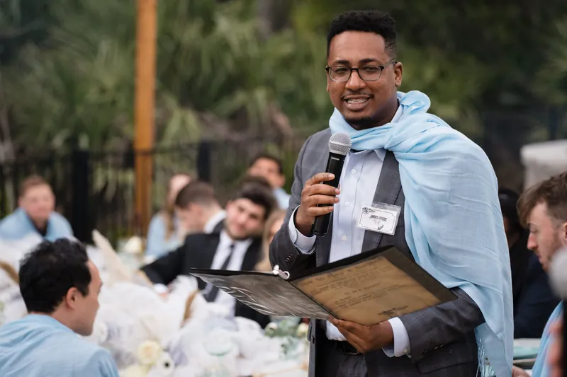Speaker in light blue shawl holding a wooden plaque and microphone, smiling at an evening reception surrounded by guests.
