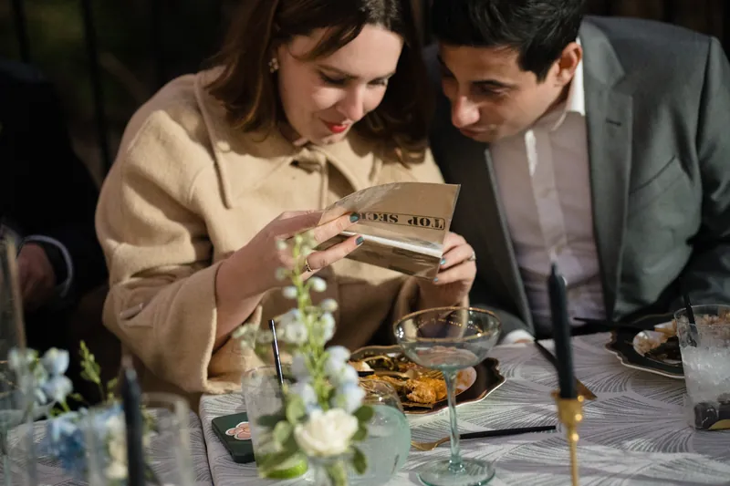 Two people at a dinner table examine a wooden plaque together, with white flowers and place settings visible.