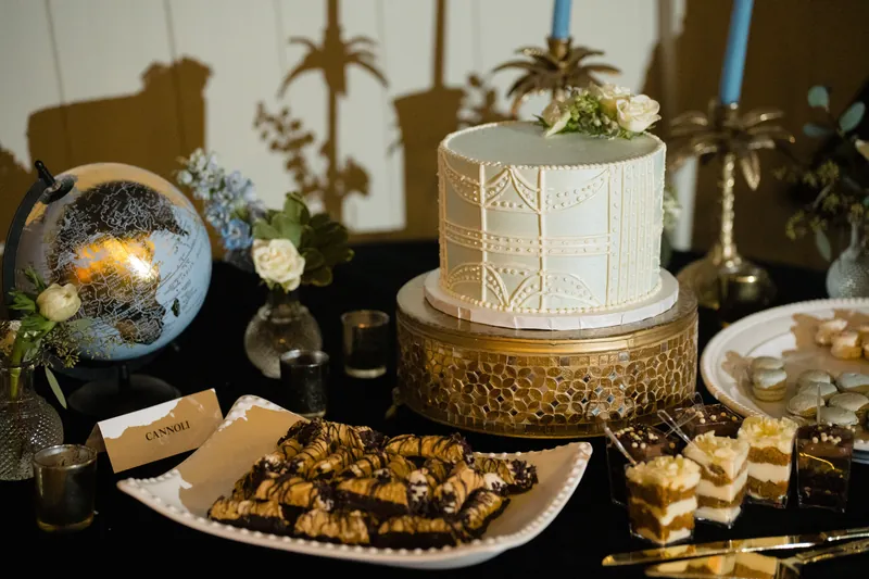 A white tiered cake with textured fondant sits on a gold stand beside golden appetizers, candied fruits, and potted succulents on a dark table.