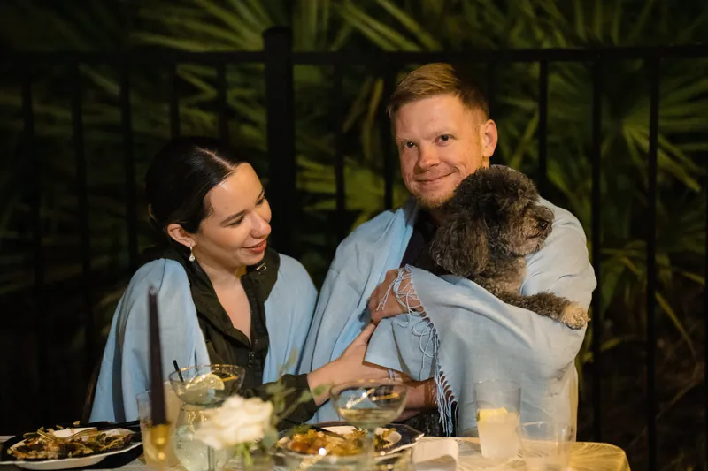 Lea and Marco sit at a candlelit dinner table, both in light blue, smiling while holding a brown dog between them.