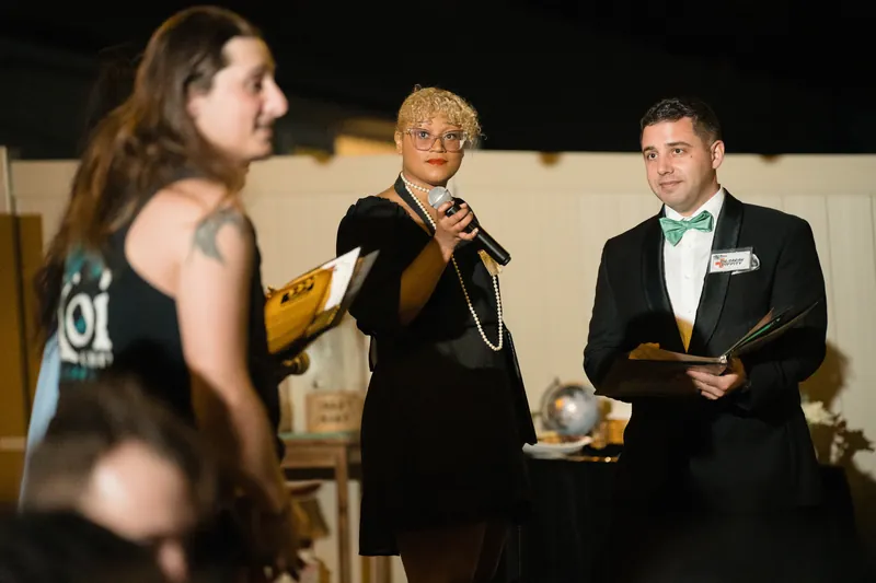 An officiant in black speaks while holding a program, with two guests in formal attire flanking them at a nighttime ceremony.