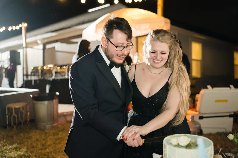 Marco in a tuxedo and a guest in a black dress cut the white cake together, surrounded by table settings and overhead lighting.
