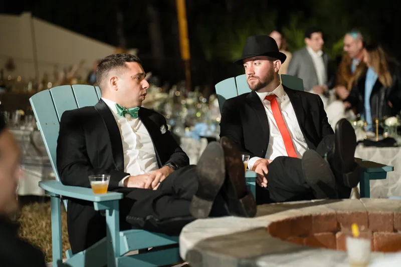 Two guests in dark suits with bow ties relax in turquoise Adirondack chairs during the evening reception.