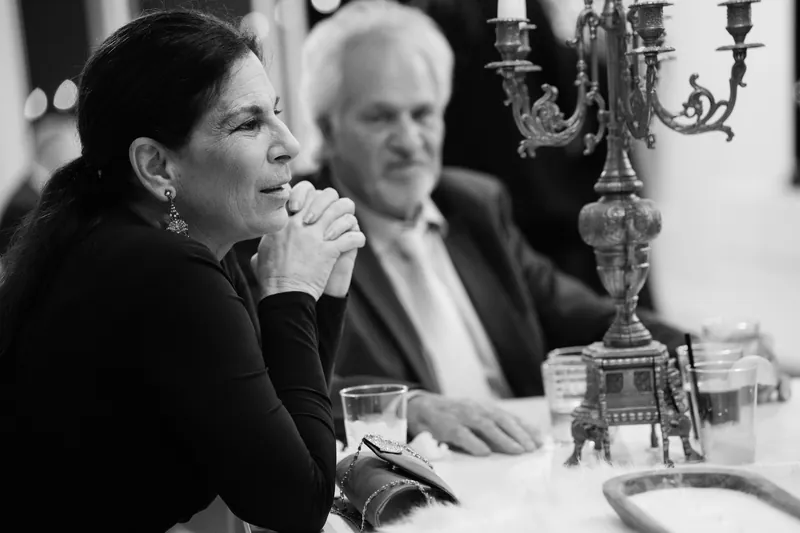 A woman in dark formal wear smiles while seated at a table set with candlesticks and glassware, a man visible beside her.