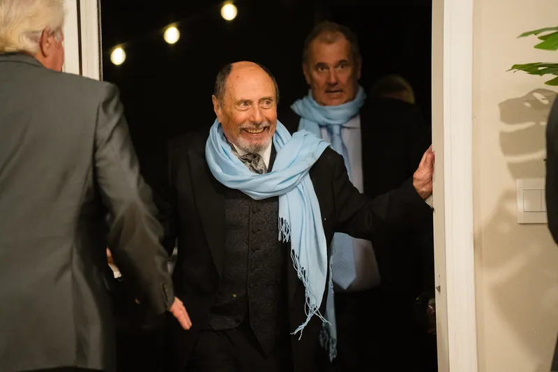 A man in a dark suit and light blue scarf stands among guests near a doorway with warm overhead lighting.