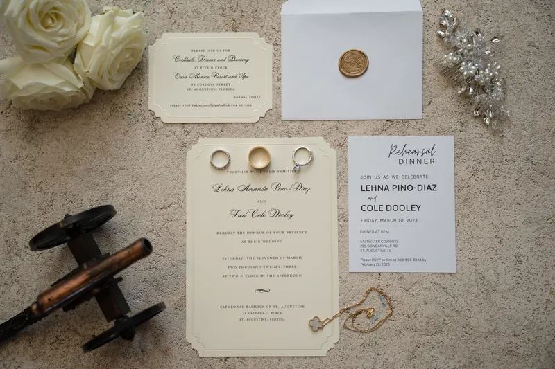 Wedding invitation suite with cream-colored cards, gold rings, twine, and white roses arranged on a neutral surface.