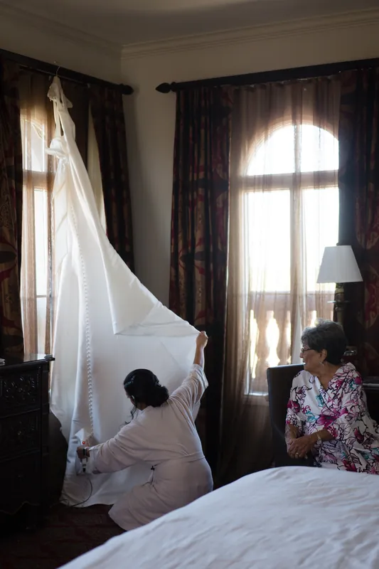 A person in a white dress stands beneath sheer fabric hanging from a window as another person adjusts the veil in a hotel