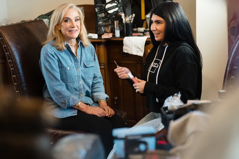 Two women chat and laugh together in a getting-ready room filled with wedding preparations and supplies.