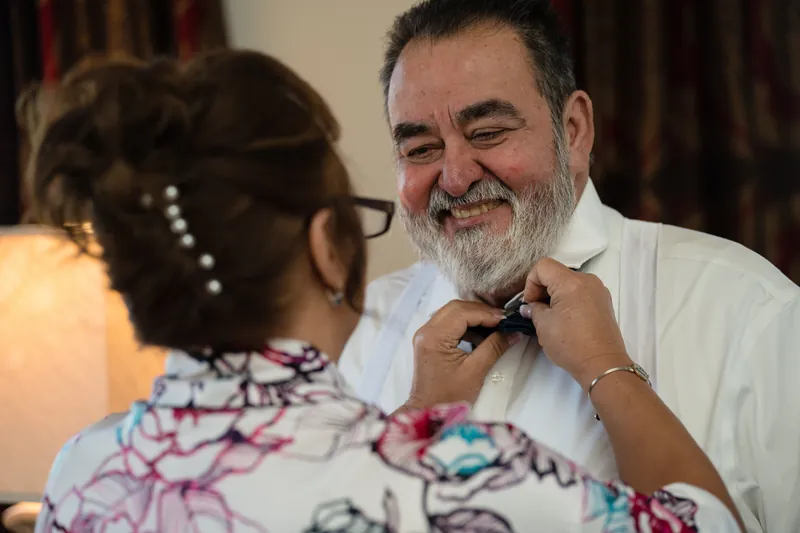 A man in a white shirt smiles while someone adjusts his bow tie before the ceremony.