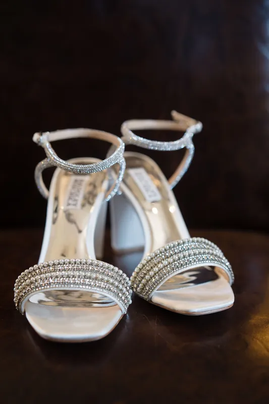 Jeweled white heeled sandals sit on a dark surface, ready for the wedding day.