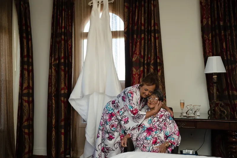 Lehna's white wedding dress hangs from a window as two women in matching floral robes help with final preparations.
