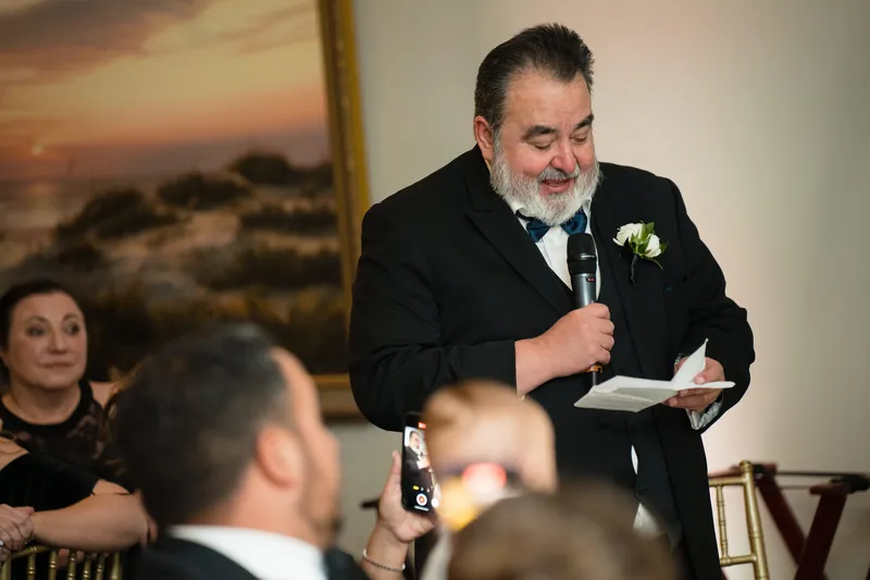 Cole speaks into a microphone at a dinner table, holding notes during his toast.
