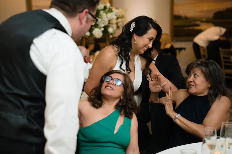 Guests laugh while one woman wearing sunglasses reacts animatedly at the reception table.