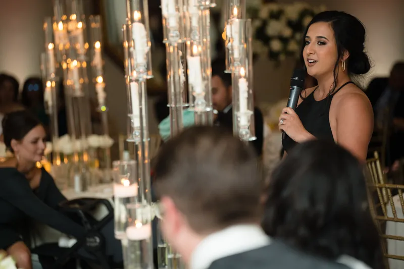 A woman in black holds a microphone and speaks to seated guests near tall brass candleholders at the reception.