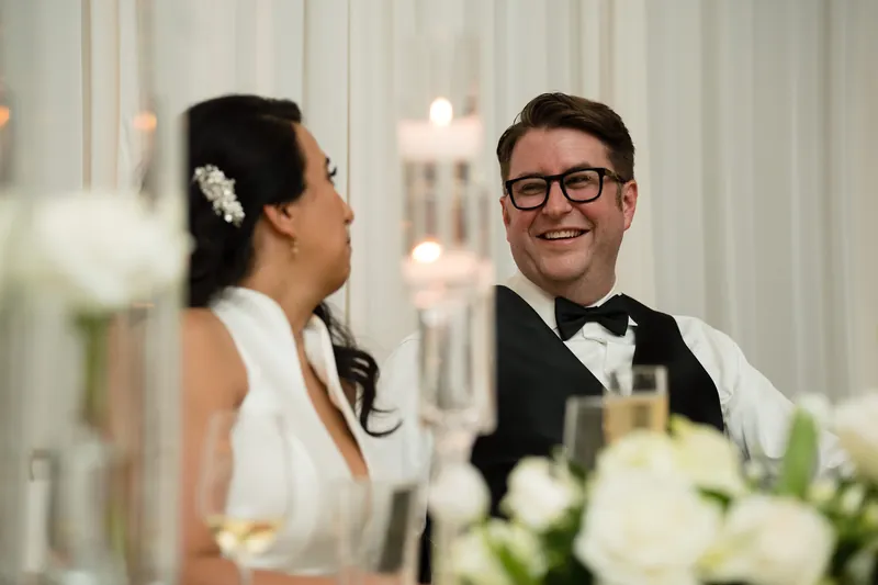 Lehna turns to smile at Cole during dinner, white flowers visible in soft focus beside them.