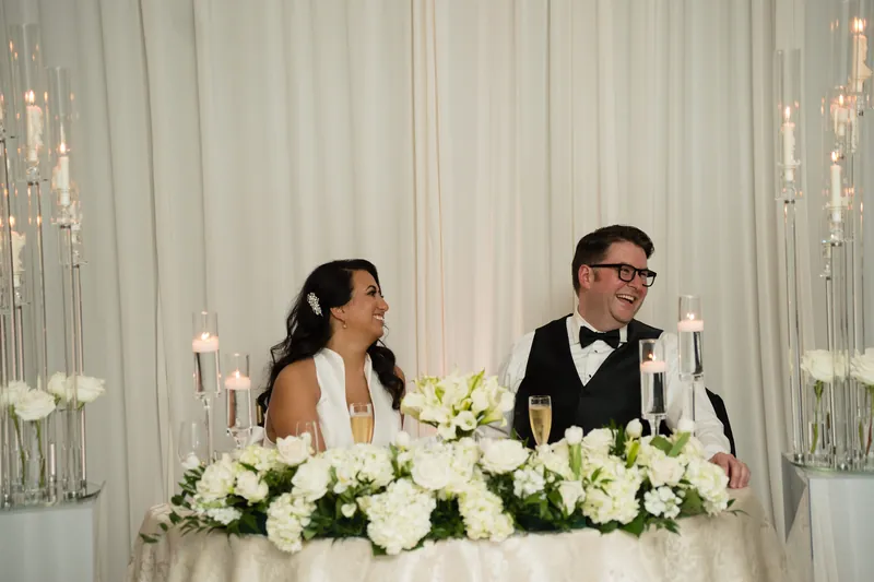 The couple sits at their head table with white floral centerpieces and tall candle arrangements flanking them.