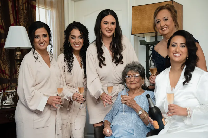 Lehna poses with family members and friends in white robes, holding champagne flutes in an elegant room.