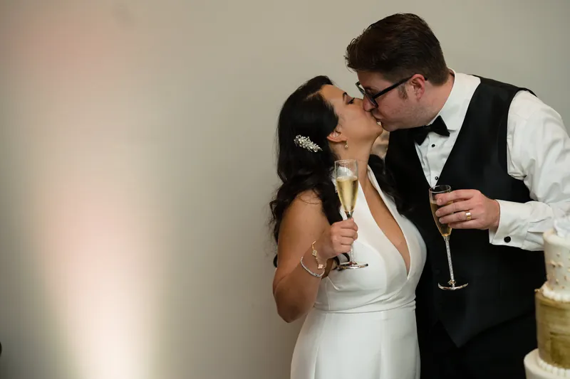 Cole kisses Lehna's cheek as she smiles, both holding champagne flutes indoors.