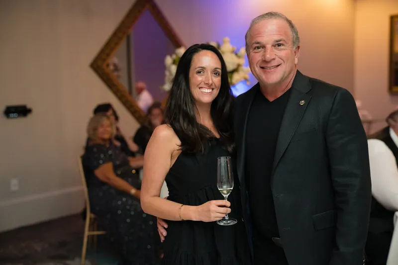 Lehna and an older man in a dark jacket smile while holding champagne glasses at the reception.