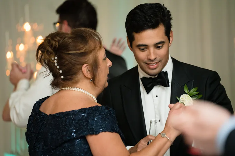Cole in a black tuxedo with bow tie and boutonniere smiles while dancing with a guest in a sequined navy dress.