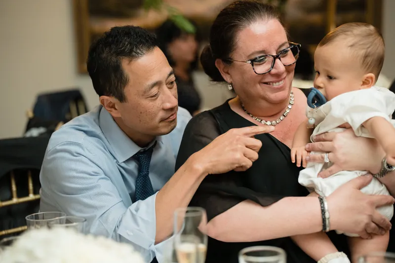 Two guests beam while holding a toddler in a white outfit during the reception.