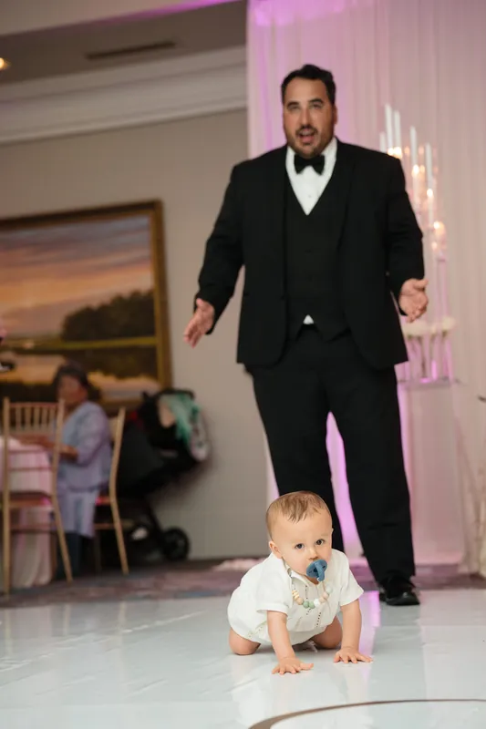 Cole in a formal black tuxedo crawls playfully across the dance floor toward a baby in a light blue outfit.