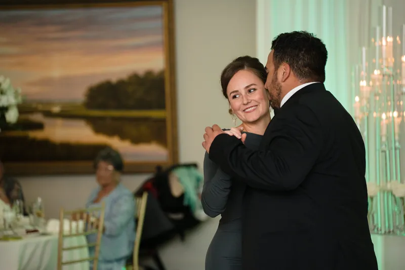 Cole embraces a woman in a black dress as they dance, with the elegant reception ballroom and artwork visible