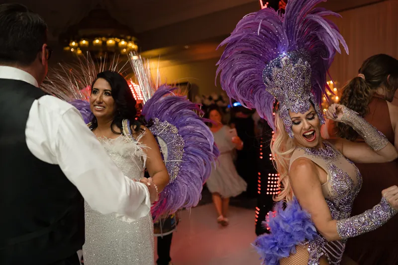 Two guests in elaborate sequined and feathered purple costumes dance with a man in formal attire at the reception.